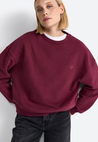 Sweatshirt oversize bordeaux avec un col rond côtelé et un logo brodé. Associé à un jean noir à coupe décontractée.