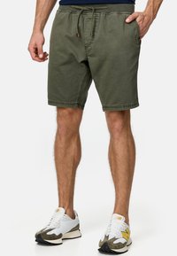 Shorts vert olive avec une taille élastique et un cordon de serrage, dotés de poches latérales et d'une coupe décontractée. Associés à des baskets blanches.