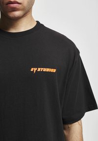 Melnas kokvilnas T-krekls ar apaļu kakla izgriezumu, ar oranžu "ZY STUDIOS" logo, kas drukāts uz kreisā krūšu laukuma. Īsas piedurknes, klasiskā piegriezums.