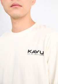 T-shirt di colore crema KAVU con maniche corte e scollatura rotonda, caratterizzato da un logo nero sul petto sinistro che recita "CHIAREZZA SOPRA VISIBILITÀ ILLIMITATA."