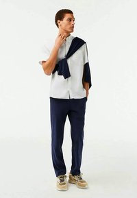 Giovane uomo in camicia bianca a maniche corte, pantaloni blu navy, sneaker beige, con un maglione blu navy appoggiato sulle spalle, che guarda di lato.