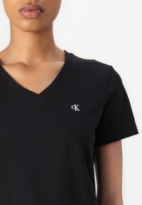 T-shirt nera a V in cotone, con un piccolo logo "ck" bianco nella zona sinistra del petto. Maniche corte e design aderente.