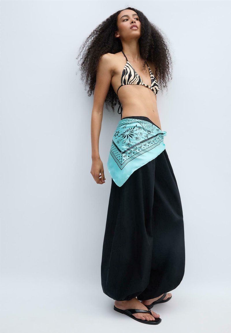 Femme aux longs cheveux bouclés portant un haut de bikini à imprimé zèbre, un pantalon noir à jambes larges, une ceinture bandana turquoise et des tongs noires.