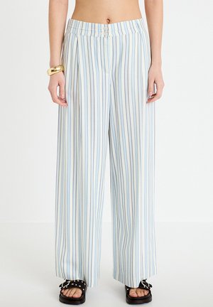 Frau trägt hoch taillierte weiße Hose mit hellblauen vertikalen Streifen, schwarze Sandalen mit Nieten und ein goldenes Armband am linken Handgelenk.