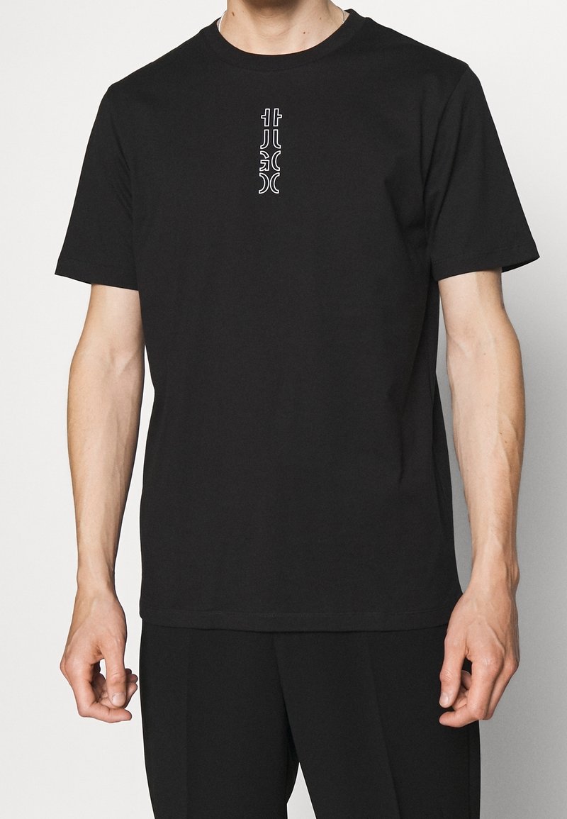 T-shirt noir à manches courtes en coton, avec un design de logo blanc vertical au centre. Encolure ras du cou simple et coupe décontractée.