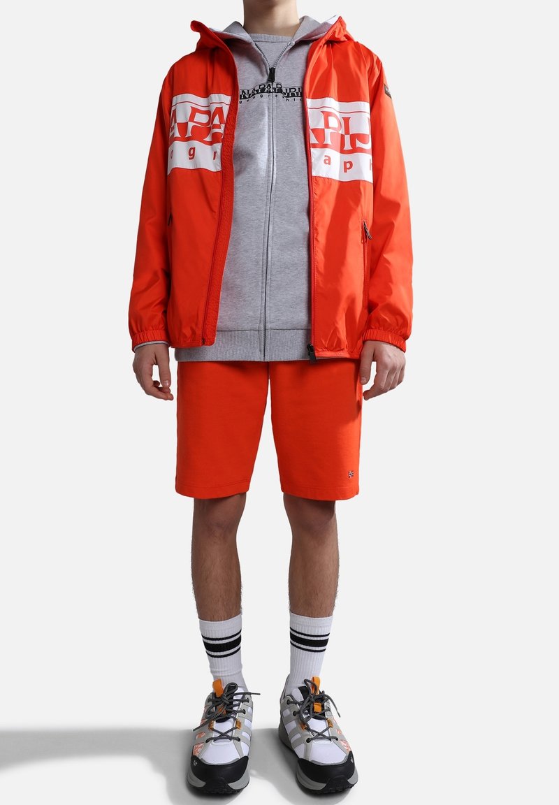 Homme portant une veste orange vif et un short, un sweat à capuche gris zippé, des chaussettes blanches avec des rayures noires, ainsi que des baskets blanches et oranges, debout sur fond blanc.