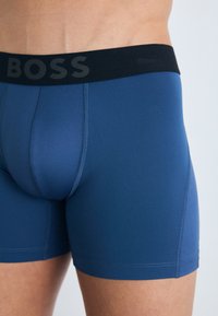 Modri bokseric z black elastičnim pasom, na katerem je v izstopajočem tisku napis "BOSS". Gladka tkanina z prilegajočim dizajnom in kontrastnimi šivi.