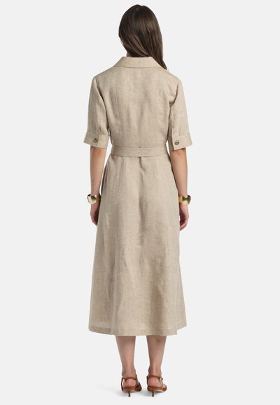 Femeie purtând o rochie bej, de lungime medie, cu curea, mâneci până la cot și detalii cu nasturi, stând cu spatele spre cameră pe un fundal alb.