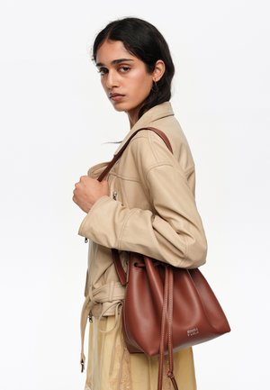 Joven con cabello oscuro que lleva una chaqueta beige y un bolso marrón de cuero con cordón sobre el hombro, sobre un fondo blanco.