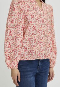 Blouse à motif floral en rose clair avec des accents rouges et dorés. Présente un col en V, des manches longues bouffantes et une coupe décontractée. Associée à un jean bleu.