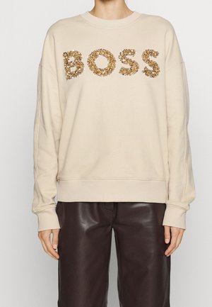 Personne portant un sweat-shirt beige avec des lettres en sequins dorés formant "BOSS" et un pantalon en cuir marron foncé, debout devant un fond uni.