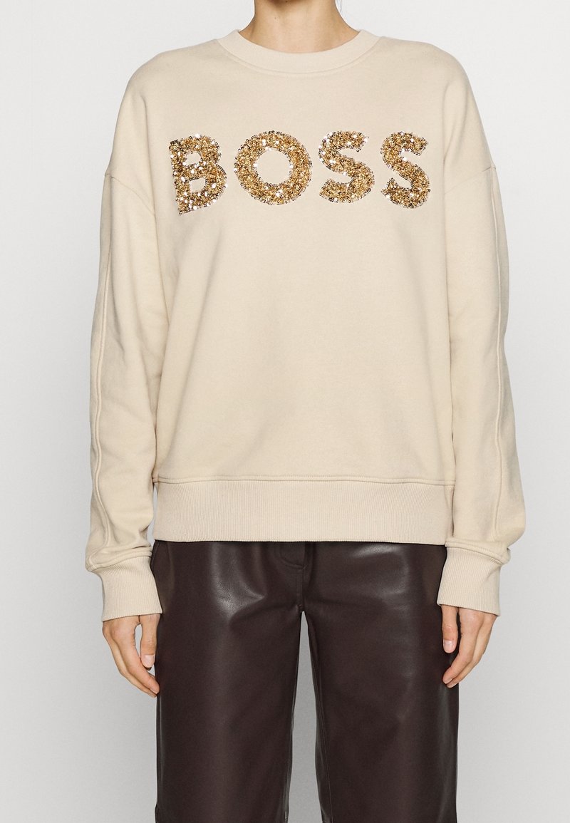 Personne portant un sweat-shirt beige avec des lettres en sequins dorés formant "BOSS" et un pantalon en cuir marron foncé, debout devant un fond uni.