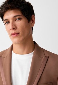 Blazer marron avec des revers crantés, texture lisse et accents aux épaules, porté par-dessus un t-shirt blanc uni. Design simple et ajusté.