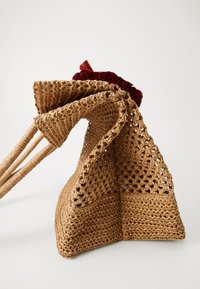 Geflochtene Strohtasche in Hellbraun mit offener Webart, einem strukturierten Boden und einem roten dekorativen Stoffakzent oben.