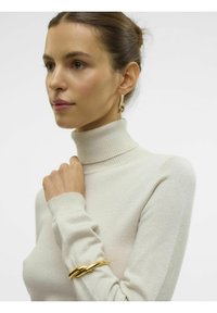 Maglione a collo alto color beige chiaro con colletto e polsini a costine, con un brillante bracciale dorato attorcigliato al polso. Texture liscia.