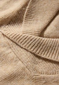 Beżowy, dziany sweter o teksturowanym, prążkowanym wzorze. Zbliżenie ukazuje miękki, przytulny charakter z nakładającymi się warstwami.