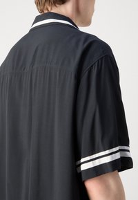 Personne portant une chemise noire à manches courtes avec des bandes blanches et noires sur les manches et le col, vue de dos et de côté.
