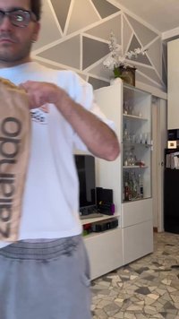 Άνδρας με γυαλιά και casual ρούχα κρατά μια καφέ χάρτινη σακούλα σε ένα μοντέρνο σαλόνι με γεωμετρική τοιχογραφία και μια γυάλινη βιτρίνα.
