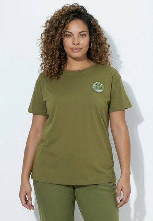 Frau mit lockigem Haar, die ein olivgrünes T-Shirt und eine Hose trägt, mit einem kleinen runden Smiley-Patch auf der Brust des T-Shirts.