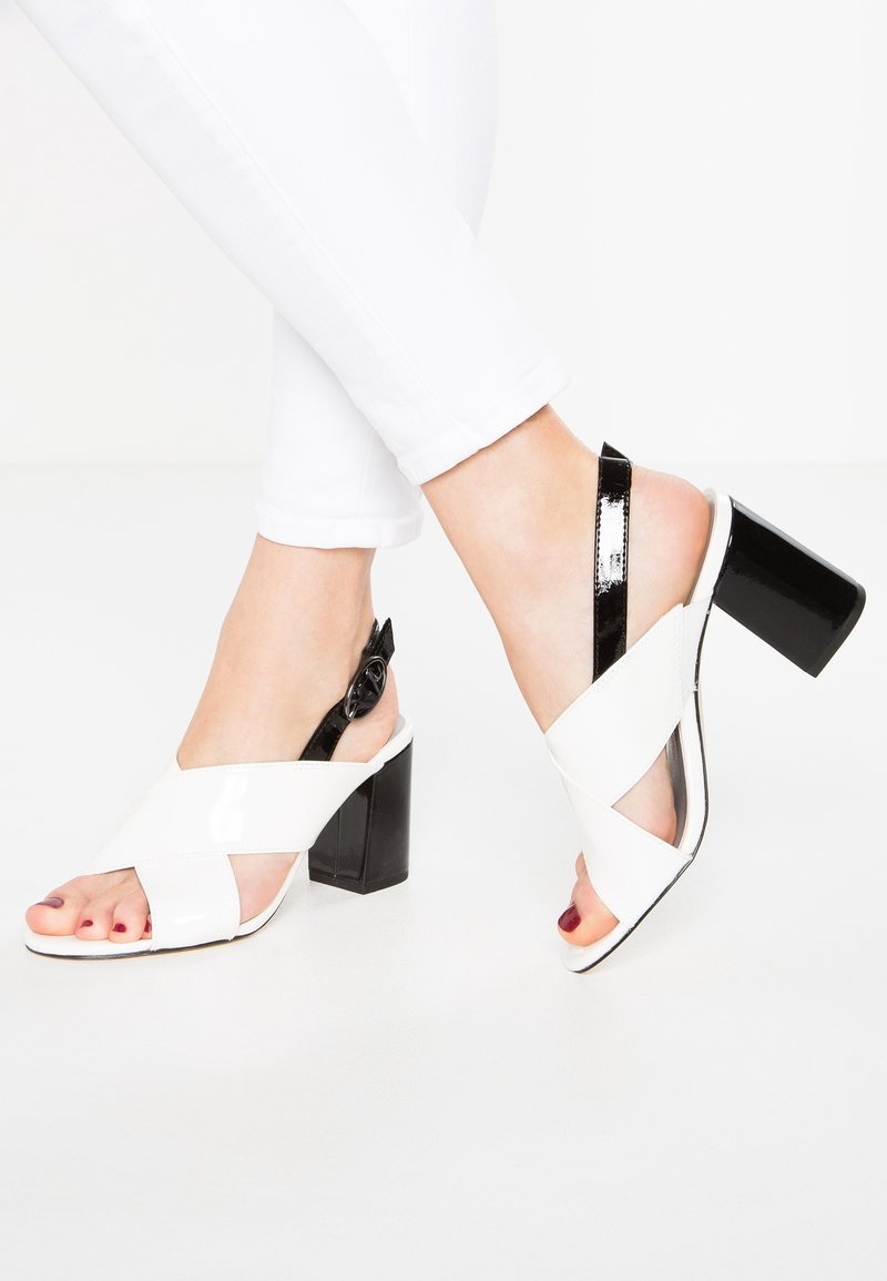 Pieds dans un pantalon blanc portant des sandales à talons ouverts blanches avec des sangles noires et des talons carrés sur un fond blanc.