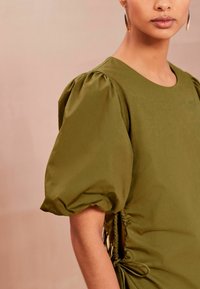 Top verde măsliniu, cu mâneci scurte și volane, decolteu rotund și detaliu de strângere pe lateral, fabricat dintr-un material fin și ușor.
