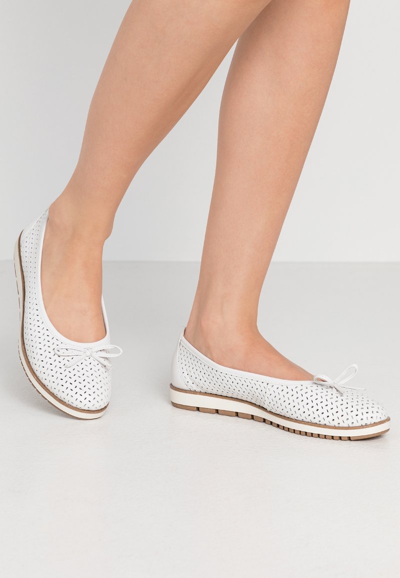 Jambes portant des ballerines blanches perforées avec de petits nœuds et des semelles en caoutchouc beige sur un sol et un fond gris clair.