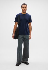 Homem vestindo uma t-shirt azul-marinho, jeans cinzentos largos, sapatos pretos, carregando uma mala de ombro preta contra um fundo claro e liso.
