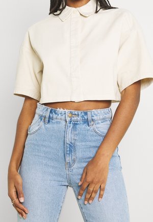 Personne portant une chemise beige courte à col et manches courtes, ainsi qu'un jean en denim bleu clair taille haute, les mains détendues le long du corps.