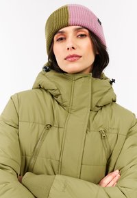 Žena s dvoubarevnou olivově zelenou a růžovou pletenou čepicí a olivově zelenou péřovou bundou s vysokým límcem a zkříženýma rukama.