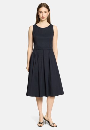 Kvinde iført sort midilængde kjole uden ærmer med plisseret nederdel og sorte højhælede sandaler med stropper, stående mod en hvid baggrund.