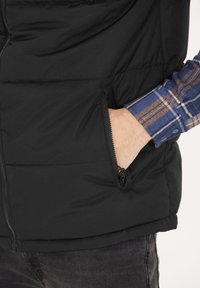 Schwarze Steppweste mit einer Reißverschlusstasche und horizontaler Steppung, kombiniert mit einem karierten Langarmhemd darunter.
