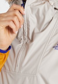 Ljusgrå jacka med en orange manschett, med dragkedja fram och en logotyp i lila. Slät textur, minimalistisk design och praktiska detaljer.