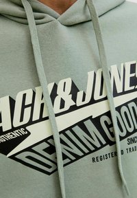 Un sweat à capuche vert avec une texture douce, présentant un grand texte graphique noir et crème indiquant "Jack & Jones Denim Goods". Cordon à la capuche.
