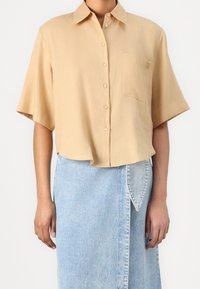 Chemise beige clair à manches courtes avec col et boutons, dotée d'une poche poitrine et d'une broderie logo discrète, associée à une jupe en denim.