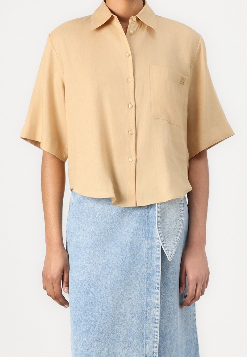 Chemise beige clair à manches courtes avec col et boutons, dotée d'une poche poitrine et d'une broderie logo discrète, associée à une jupe en denim.