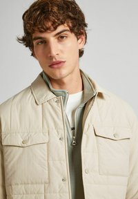 Beige quilted jacket with a pointed collar, featuring two chest pockets and a zippered inner layer in light green. Smooth texture, lightweight design.