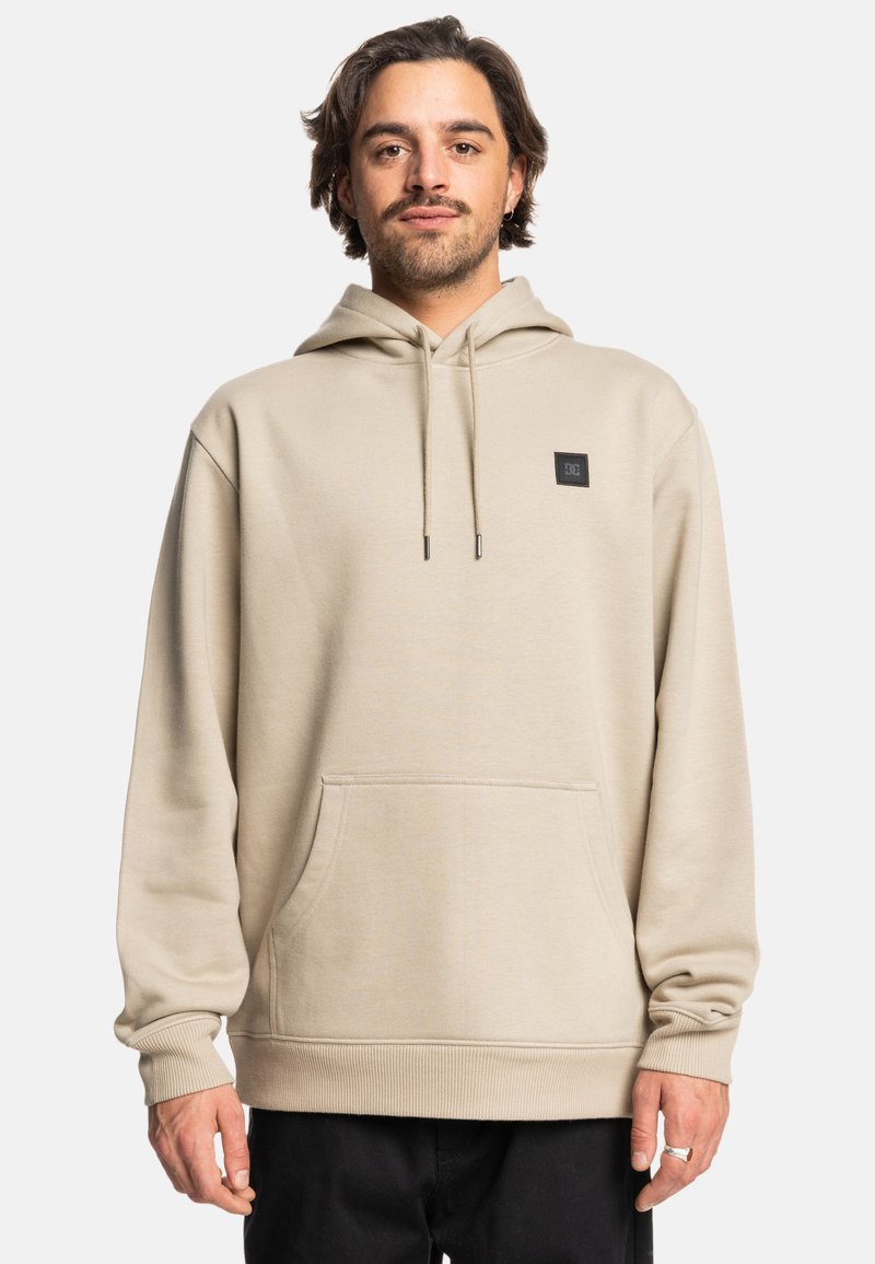 Hoodie de color beige claro con un bolsillo de bolsillo frontal, capucha con cordón y un parche de logo negro en el pecho. Tejido suave y texturizado, corte relajado.