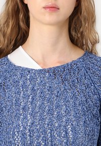 Top tricoté bleu clair avec un motif ajouré, col rond et manches courtes bouffantes, superposé à une chemise blanche à col.