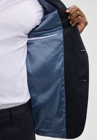 Donkerblauwe blazer met een gestructureerd patroon, voorzien van een lichtblauw satijnen interieur en een zak met een witte rand.