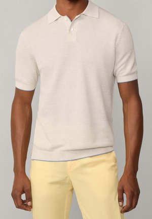 Homme portant un polo en maille beige à manches courtes associé à un pantalon jaune clair, debout devant un fond neutre.