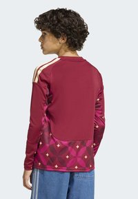 Maroon athletic long-sleeve shirt with cream stripes on shoulders and a patterned lower section in shades of pink and decorative shapes.