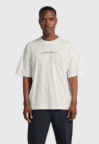 Vit bomulls-t-shirt med oversize passform, rund halsringning och korta ärmar. Har svart tryckt text på bröstet.