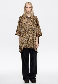 Top de estampado de leopardo con capucha ajustable, corte oversize, mangas cortas y diseño en capas, combinado con pantalones negros holgados y zapatos oscuros.