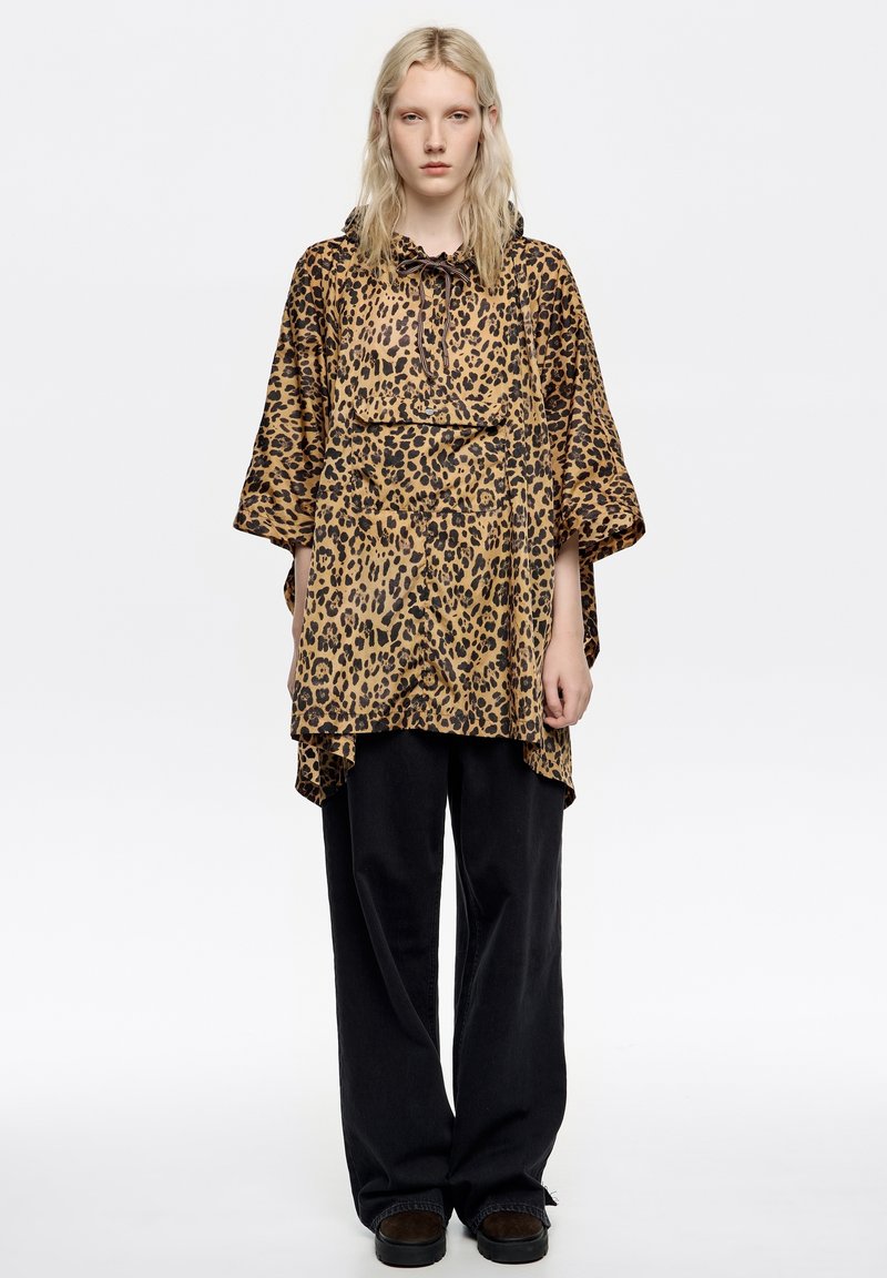 Top de estampado de leopardo con capucha ajustable, corte oversize, mangas cortas y diseño en capas, combinado con pantalones negros holgados y zapatos oscuros.