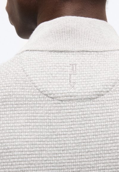 Κοντινό πλάνο του λαιμού και του άνω μέρους της πλάτης ενός ατόμου που φοράει ένα υφασμάτινο λευκό πουλόβερ με ανάγλυφο λογότυπο "JUX" στο γιακά.