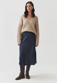 Beige gebreide trui met V-hals, losse pasvorm, gecombineerd met een marineblauwe satijnen midi-jurk. Bruine laarzen tot de knie maken de outfit compleet.