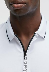 Polo shirt blanc avec une fermeture éclair noire et un col rehaussé, arborant un design sportif et un détail de tirette de fermeture éclair avec logo. Texture du tissu lisse visible.