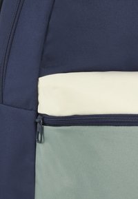 Zaino in tessuto blu navy e verde chiaro con un pannello crema, dotato di una tasca con cerniera e superficie testurizzata. Costruzione resistente con linee pulite.