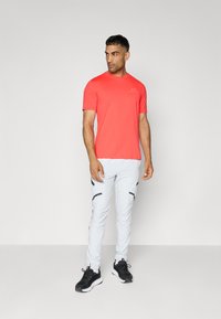 Rød idretts kortermet skjorte, grå tapered bukser med glidelåser, og svarte joggesko. Glatt stoff, minimal merking, og sporty design.