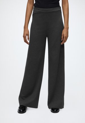 Personne portant un pantalon large gris foncé associé à des chaussures noires brillantes et un haut noir, debout devant un fond clair uni.